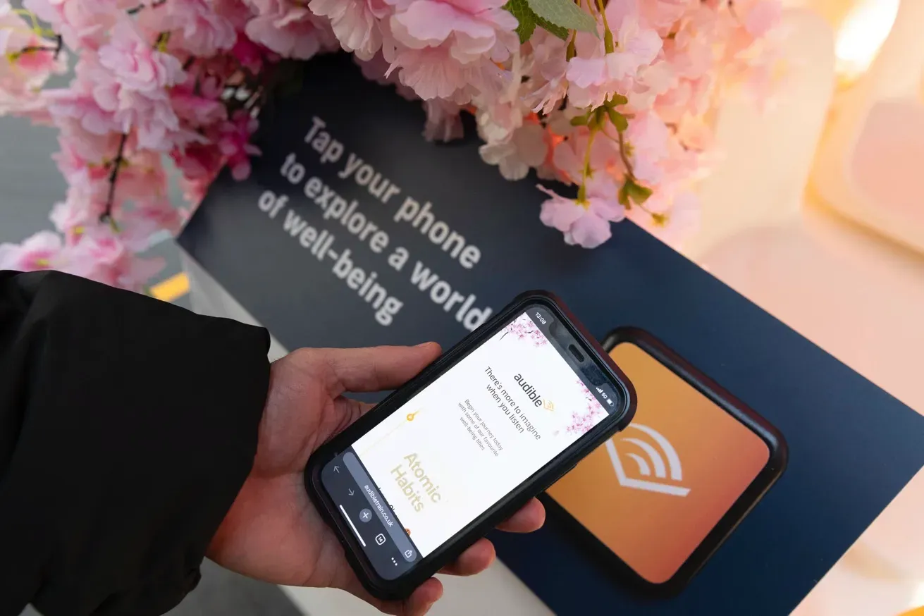 Person holding a smartphone near a sign with flowers, displaying an Audible app screen with an option to explore content related to well-being.