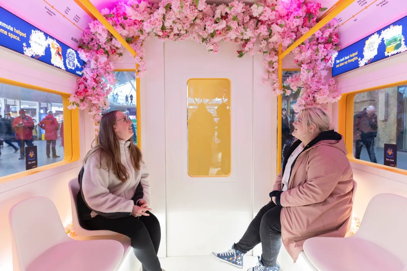 Two people sit across from each other in a brightly lit train carriage decorated with pink flowers on the ceiling.