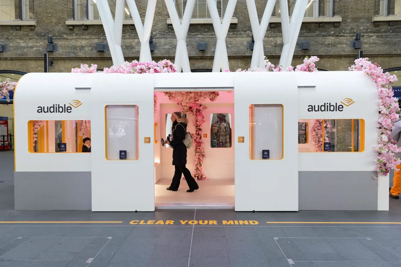 White Audible booth shaped like a train carriage, adorned with pink flowers. People are walking inside. The words "CLEAR YOUR MIND" are on the ground.