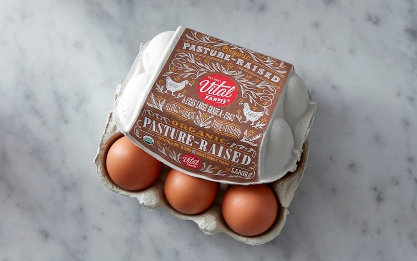 A carton of Vital Farms organic pasture-raised brown eggs on a marble surface, with two eggs visible outside the carton.