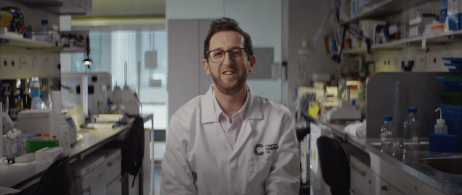 A person in a lab coat smiles while sitting in a laboratory filled with scientific equipment and supplies.