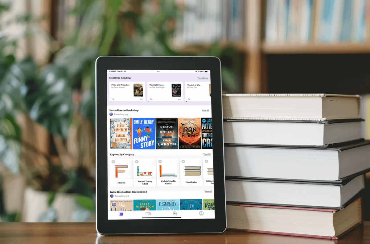 A tablet displaying a digital bookstore app stands beside a stack of hardcover books, with a bookshelf in the background.