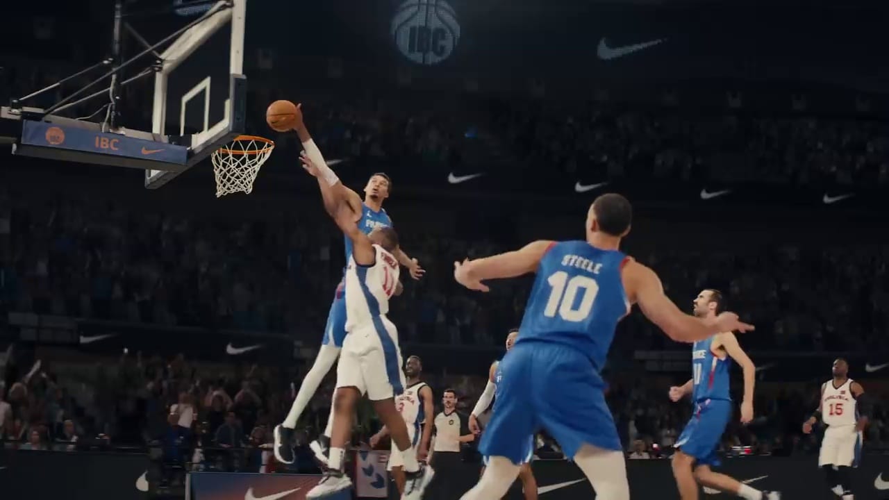 A basketball player in a blue jersey attempts to block a shot by a player in a white jersey near the hoop, while teammates look on during a game.