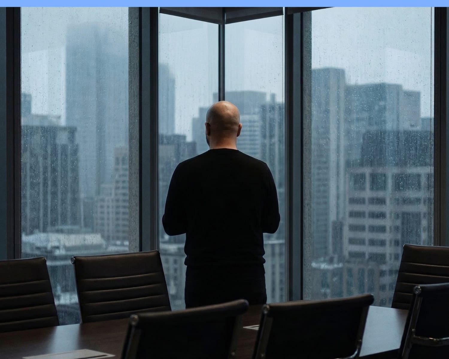 A person stands alone in a conference room, looking out large windows at a rainy city skyline with tall buildings.