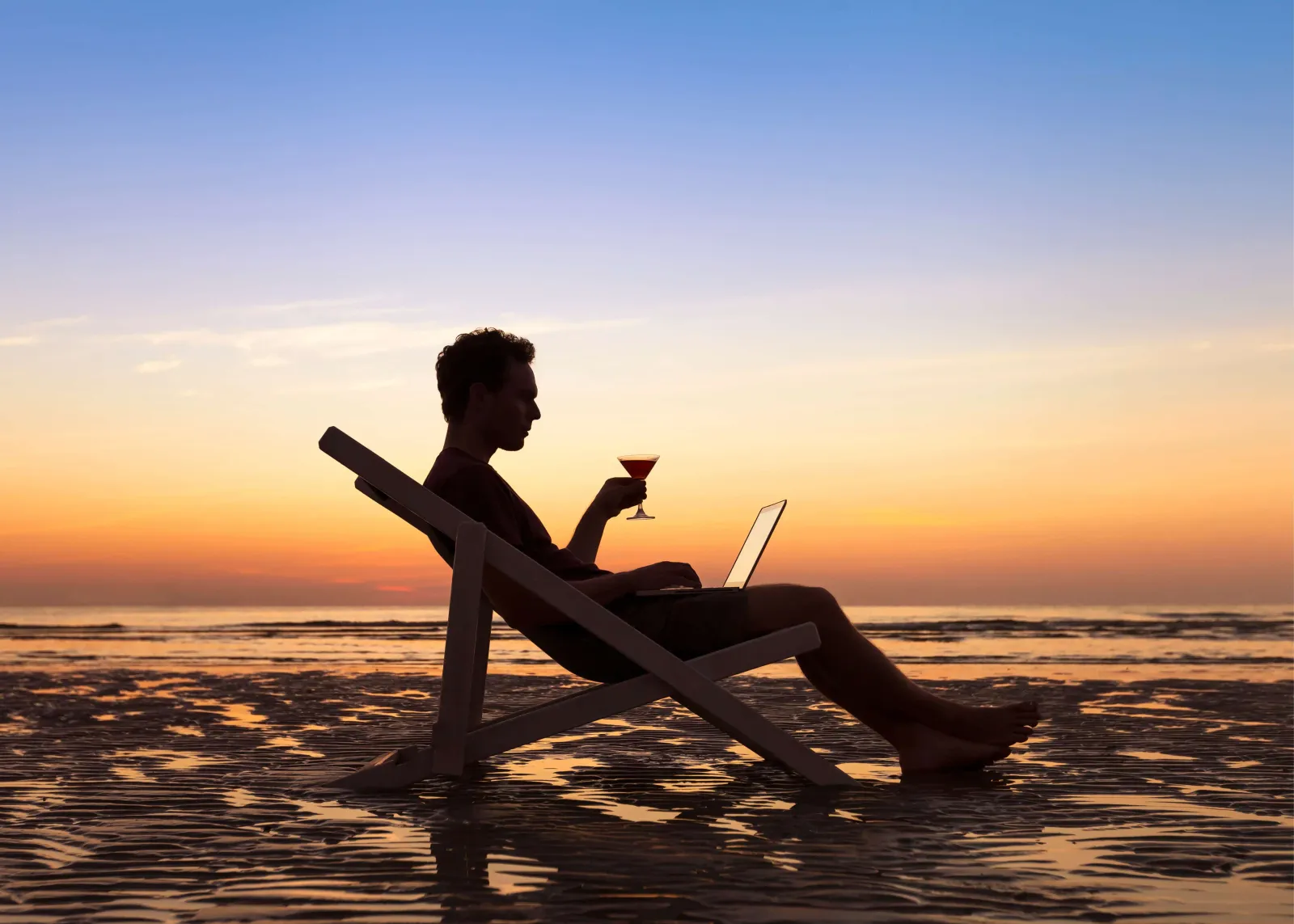 A man sat on a sun chair, on a beach during sunset with a margarita in had and laptop on leg, illustrating the ability to work from anywhere at anytime.