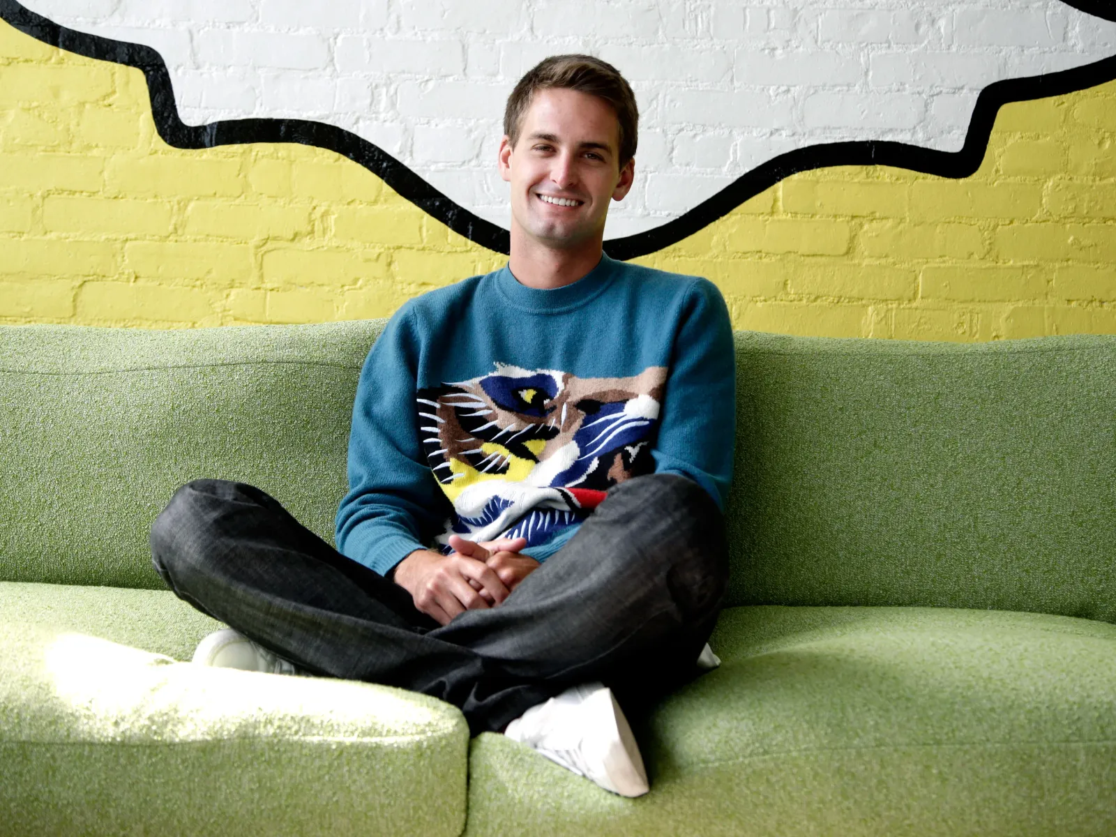 Evan Spiegel (Snapchat founder) smiling on a green couch, wearing a colorful sweater with an abstract design. Yellow and white mural background creates a playful mood.