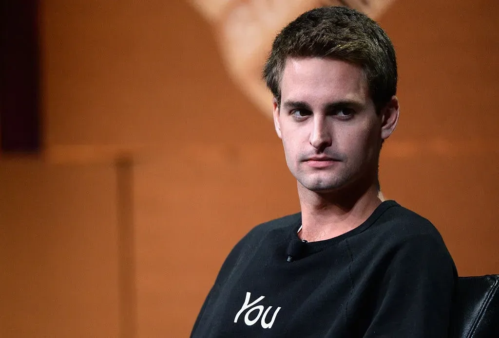 Evan Spiegel in a black sweatshirt with "You" printed on it, seated against a brown backdrop, looking to the side with a serious expression.