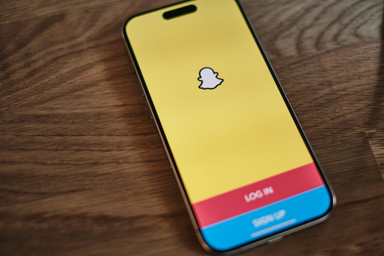 Smartphone on wooden surface displaying the Snapchat login screen. The screen has a yellow background with a white ghost icon, "LOG IN" and "SIGN UP" options.