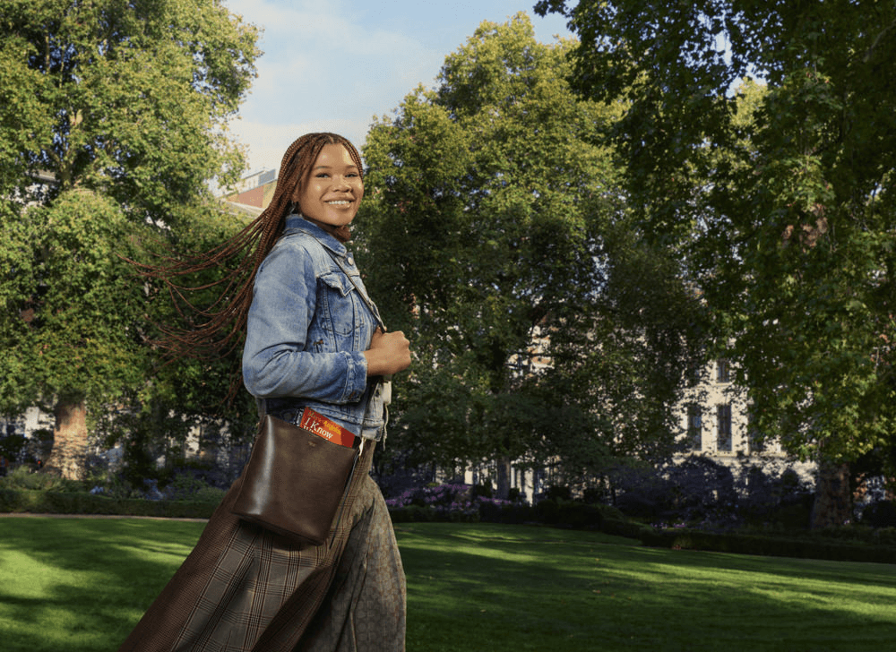 A woman in a denim jacket and plaid skirt walks through a sunny park with trees and buildings in the background, carrying a brown shoulder bag.