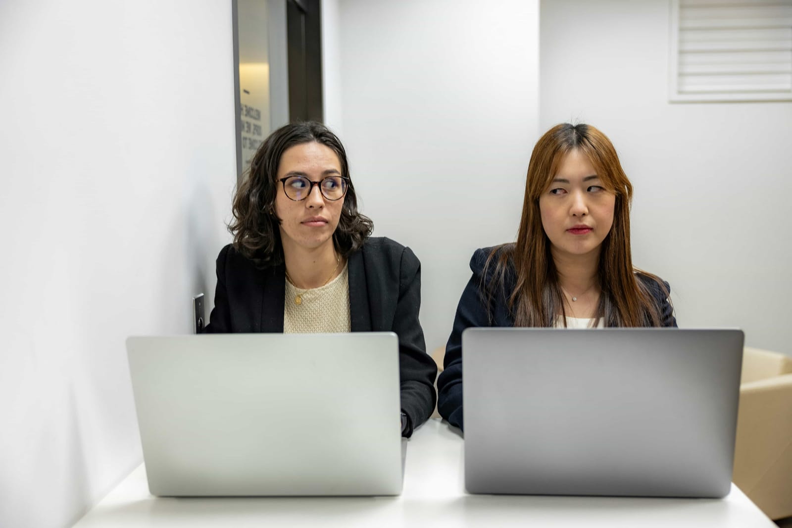 Two women sitting at a desk with laptops in front of them, seemingly unhappy at work.